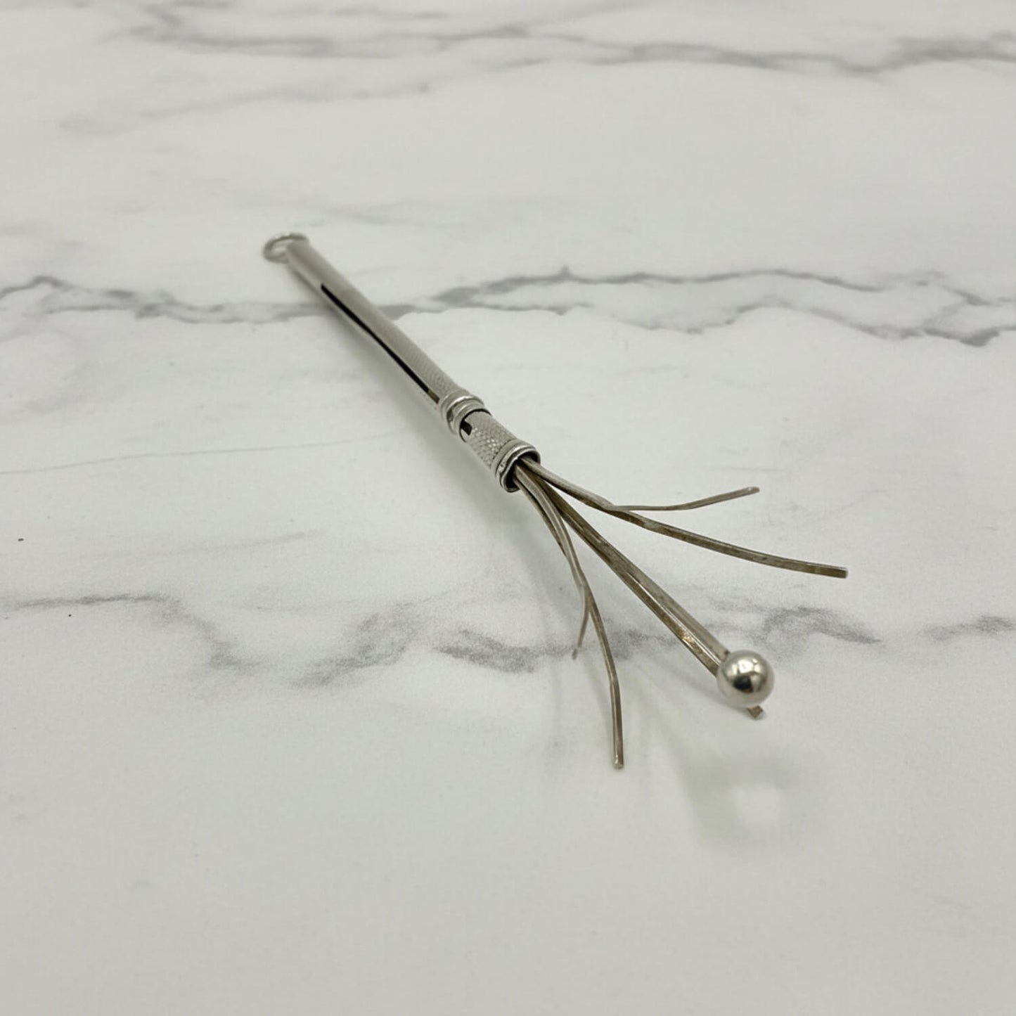 A silver Art Deco Sterling Silver Cocktail Whisk with a textured handle, a small ball tip, and several thin prong-like wires, photographed on a white marble surface.