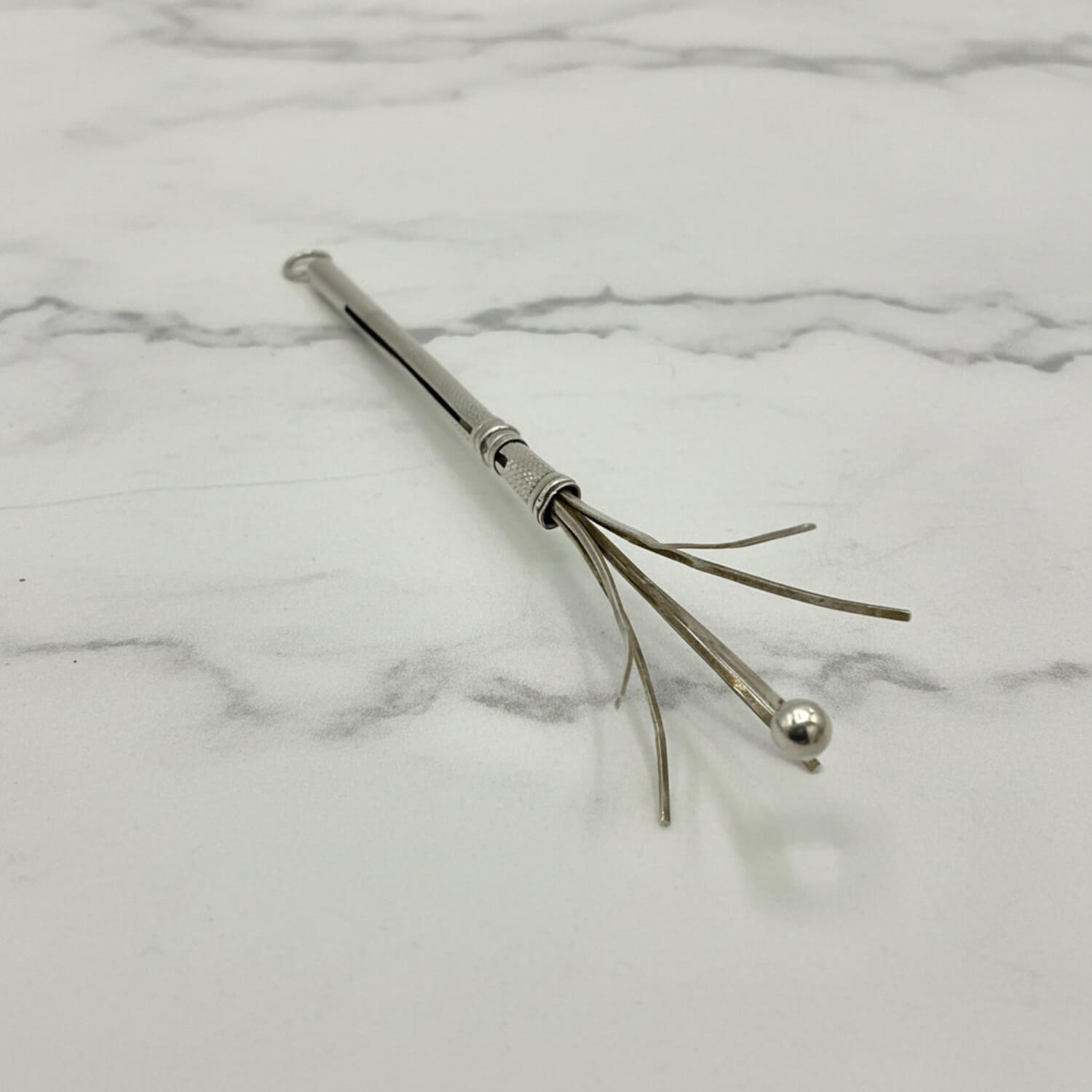 A silver Art Deco Sterling Silver Cocktail Whisk with a textured handle, a small ball tip, and several thin prong-like wires, photographed on a white marble surface.