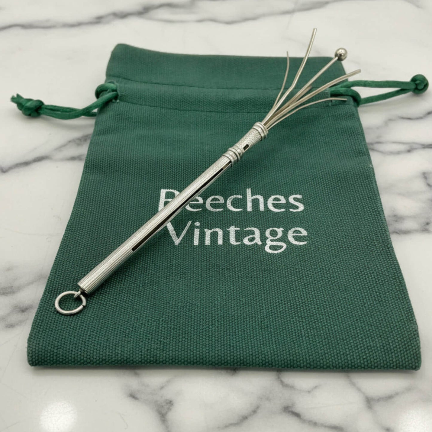 An Art Deco Sterling Silver Cocktail Whiskwith a textured handle and fine wire ends, displayed on top of a green drawstring pouch printed with 'Beeches Vintage' on a white marble surface.