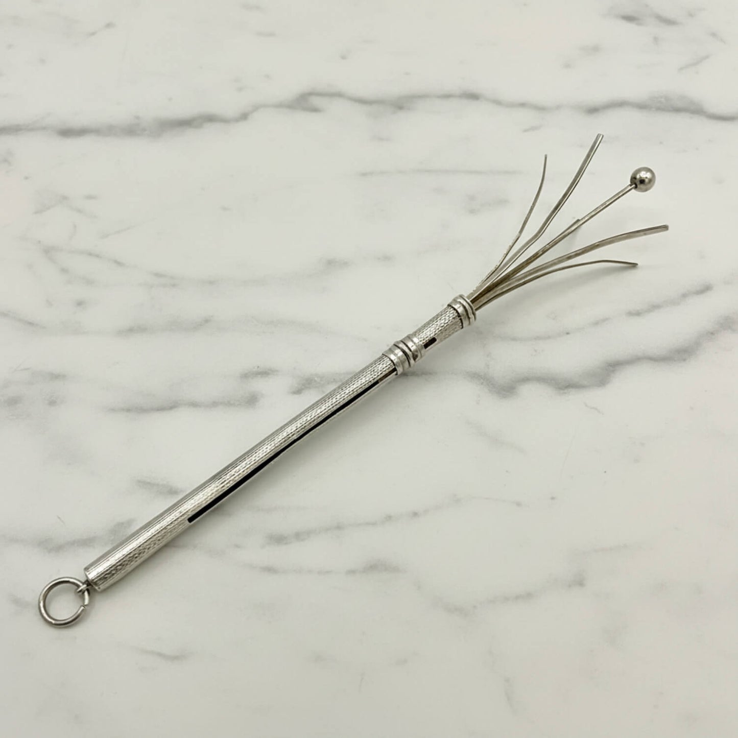 A silver metal cocktail whisk with a textured handle, a hanging loop, and fine wire bristles ending in a small ball, photographed on a white marble surface.