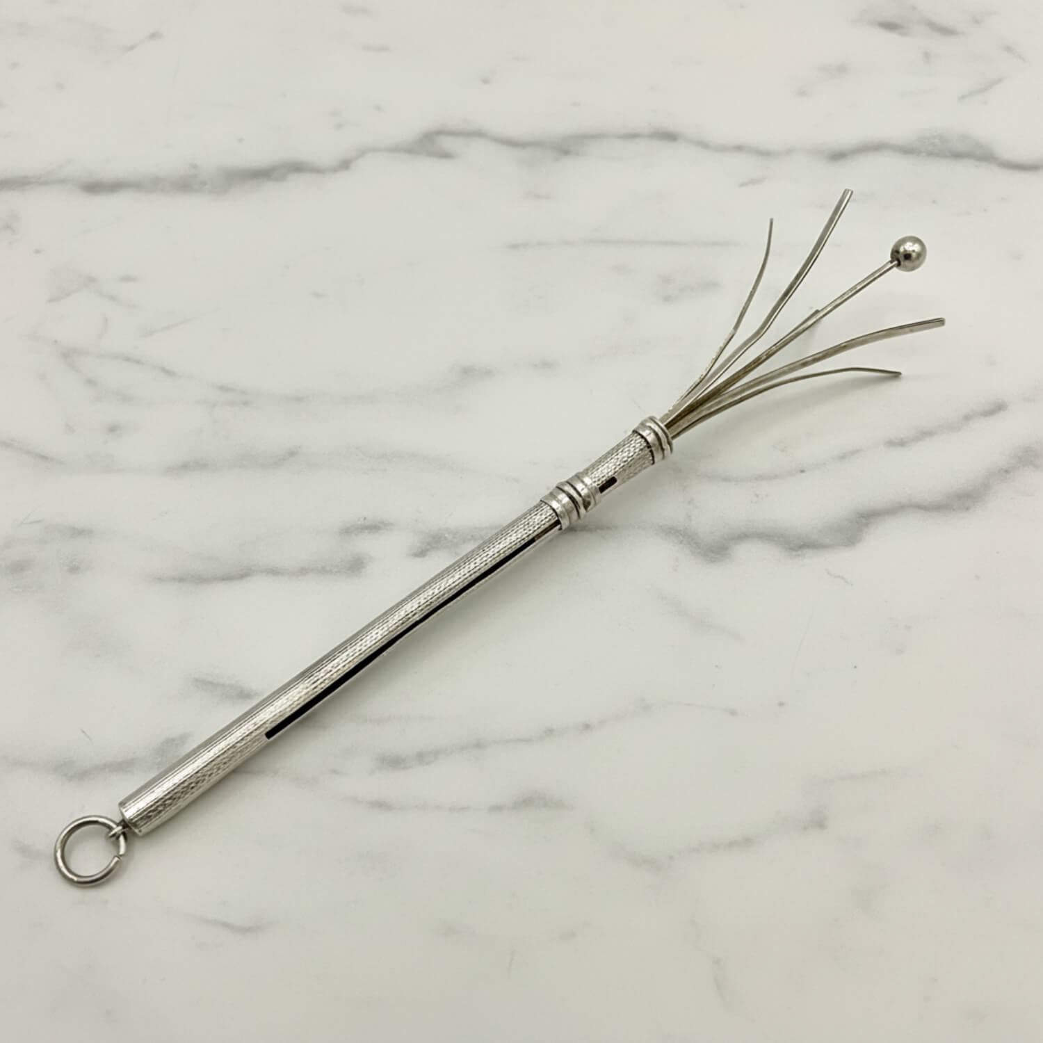 A silver metal cocktail whisk with a textured handle, a hanging loop, and fine wire bristles ending in a small ball, photographed on a white marble surface.
