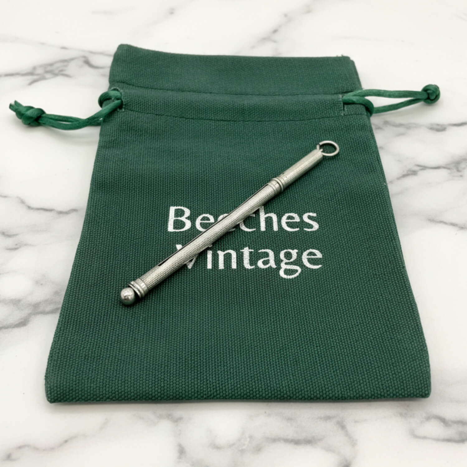 A Art Deco Sterling Silver Cocktail Whisk, displayed diagonally on a dark green drawstring pouch with 'Beeches Vintage' printed in white, set on a white marble surface.