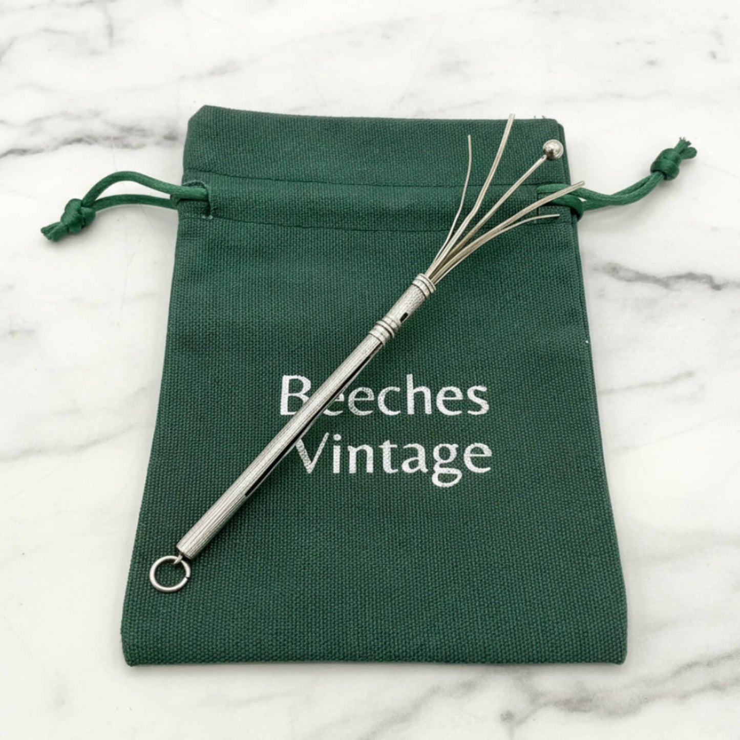 A Art Deco Sterling Silver Cocktail Whisk on a dark green drawstring pouch printed with the brand name 'Beeches Vintage', set on a white marble surface.