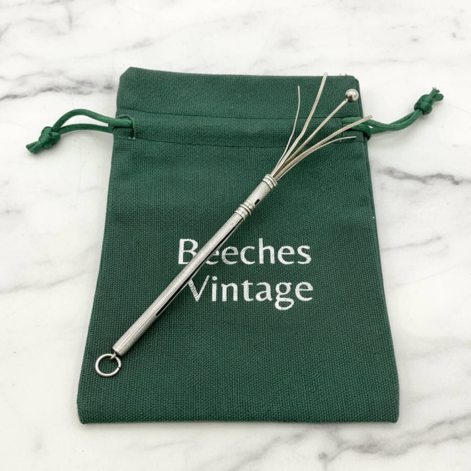 A Art Deco Sterling Silver Cocktail Whisk on a dark green drawstring pouch printed with the brand name 'Beeches Vintage', set on a white marble surface.