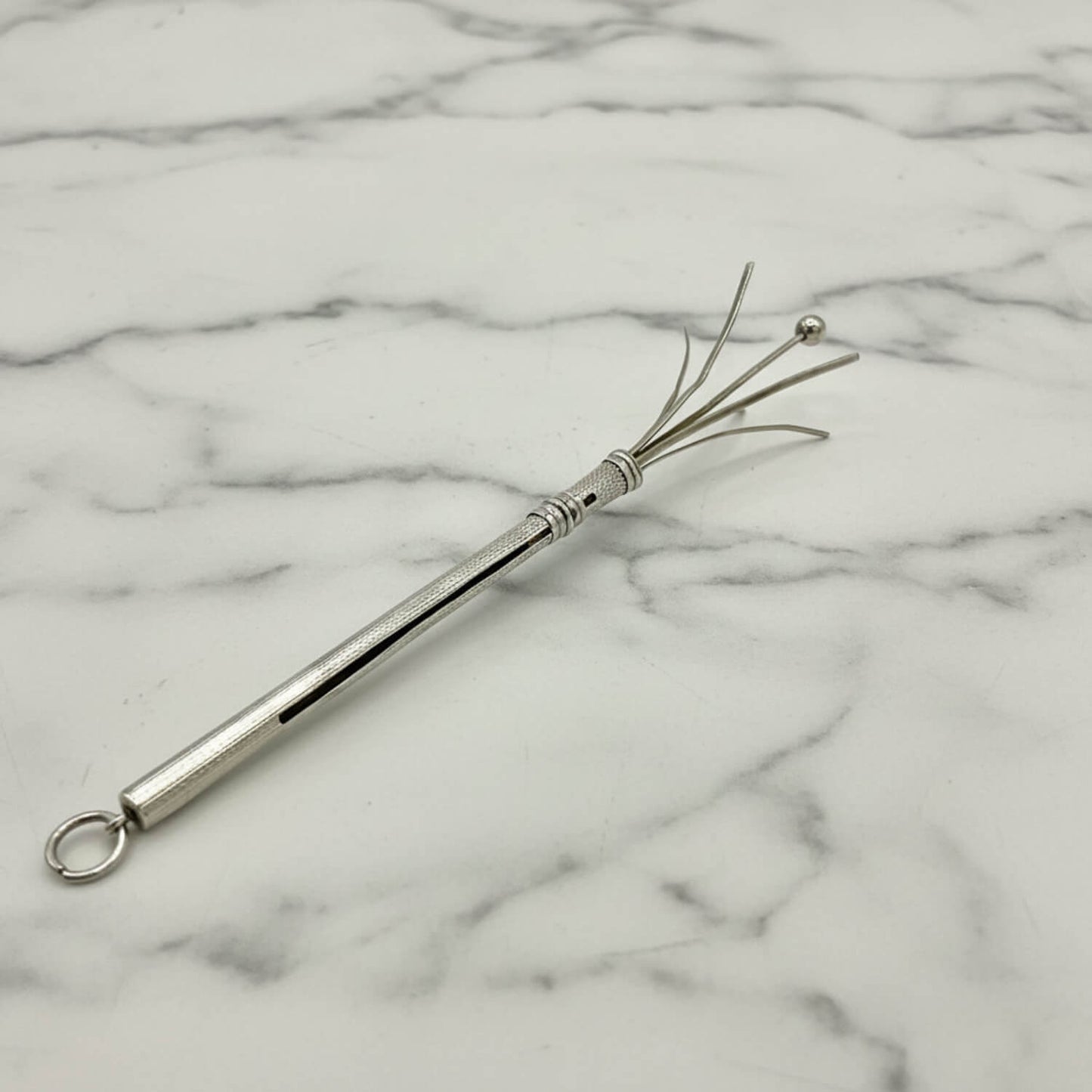 A Art Deco Sterling Silver Cocktail Whisk with a textured handle, a hanging loop, and four fine bristles ending in a small ball, displayed on a white marble surface.