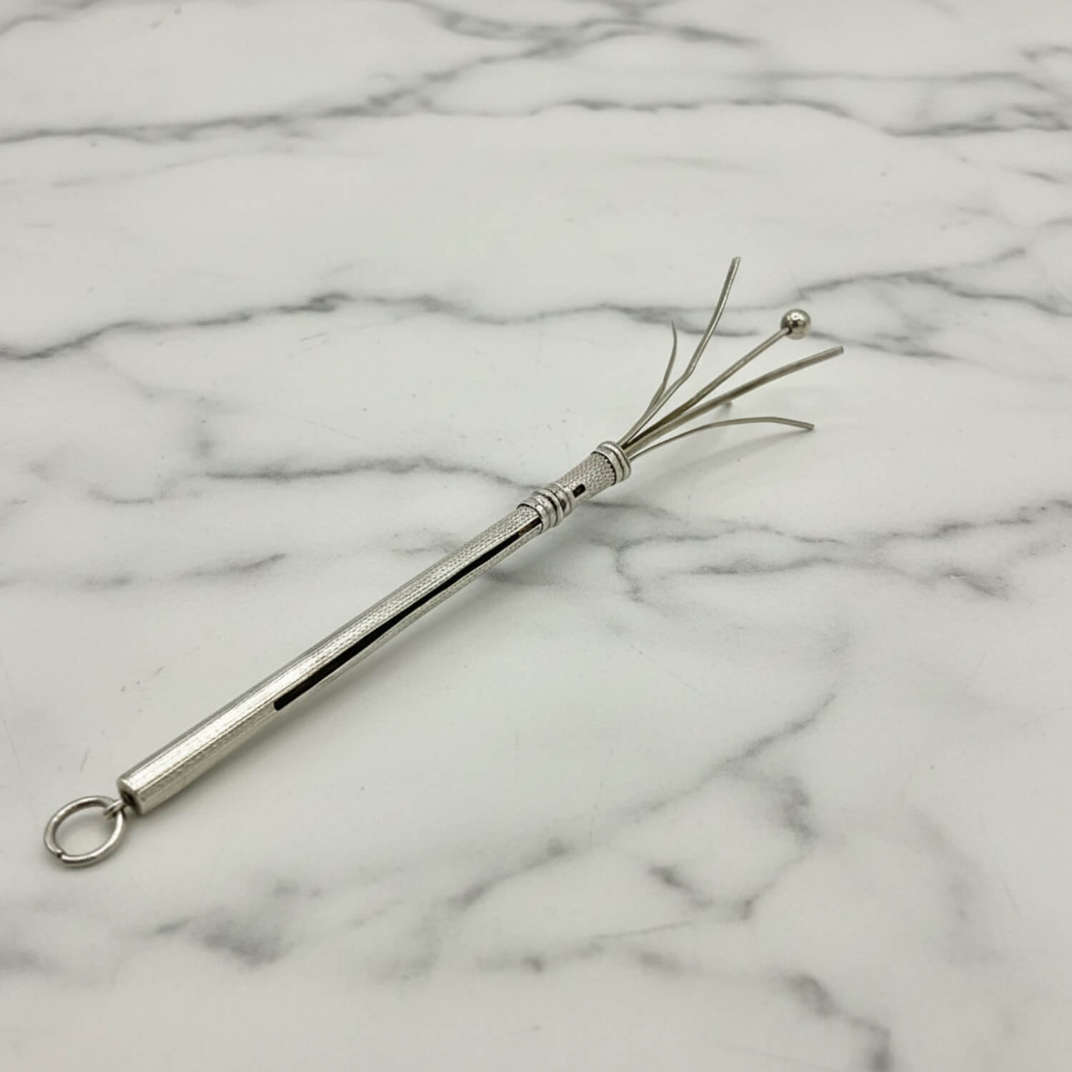 A Art Deco Sterling Silver Cocktail Whisk with a textured handle, a hanging loop, and four fine bristles ending in a small ball, displayed on a white marble surface.