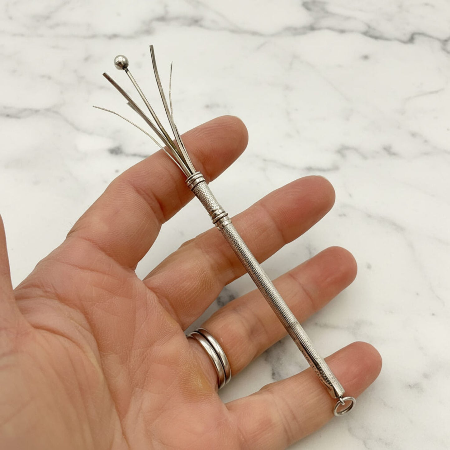 A Art Deco Sterling Silver Cocktail Whisk  with a textured handle and multiple thin prongs, held in a person's hand against a white marble background.