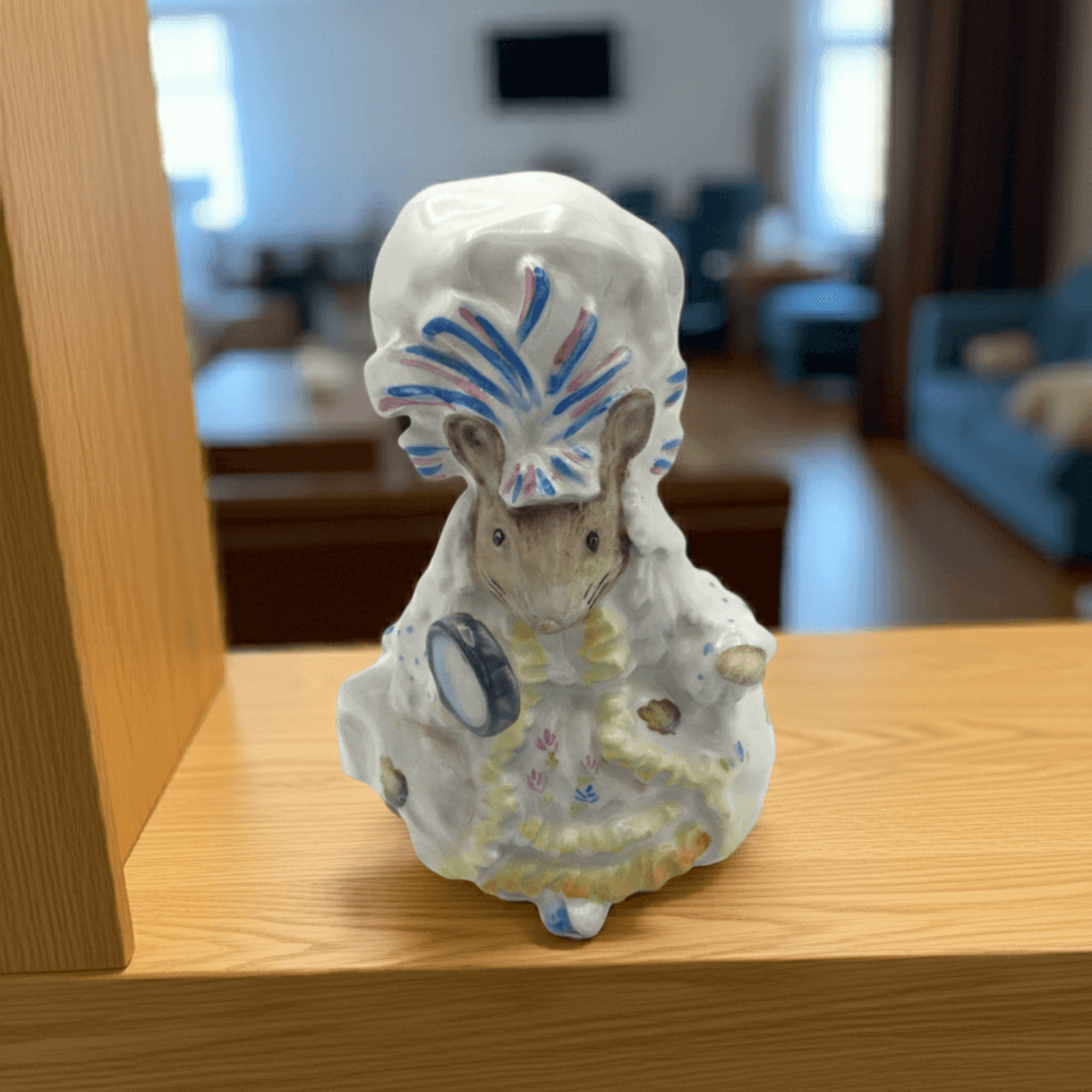 Decorative figurine of a mouse in a dress and hat on a wooden surface with a blurred indoor background.