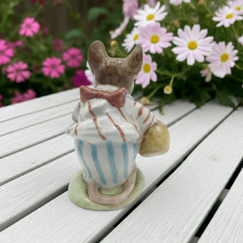 Small figurine of a mouse in striped clothes on a wooden surface with flowers in the background