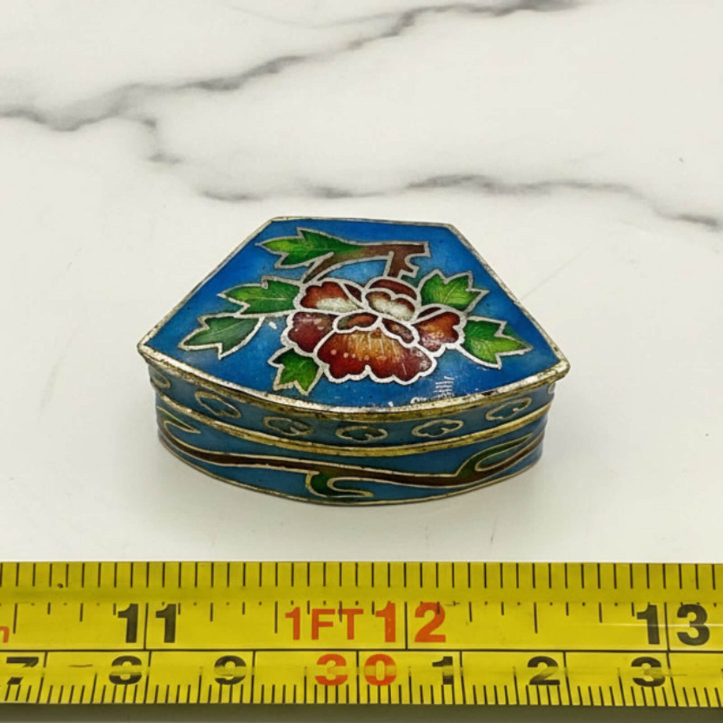 Decorative hexagonal box with floral design on a marble surface, measured against a tape measure.