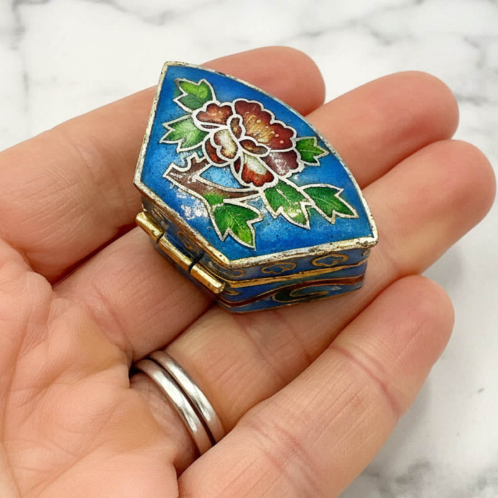 Small decorative box with floral design held in a hand against a marble background