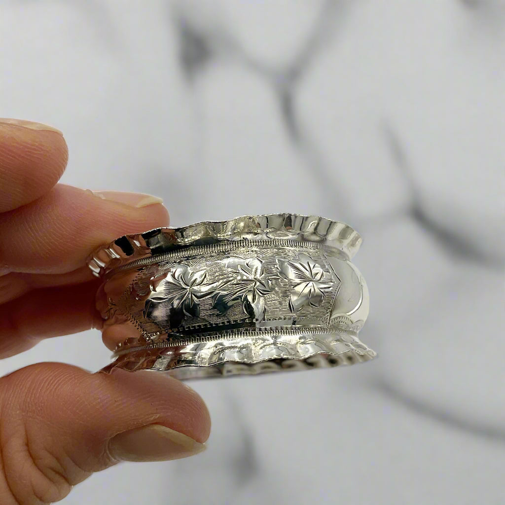 Silver napkin ring with intricate art nouveau designs held by a hand against a blurred marble background