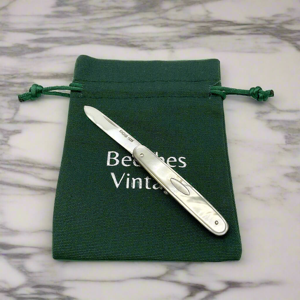 Silver pocket knife on a green drawstring bag with 'Beeches Vintage' branding on a marble surface.