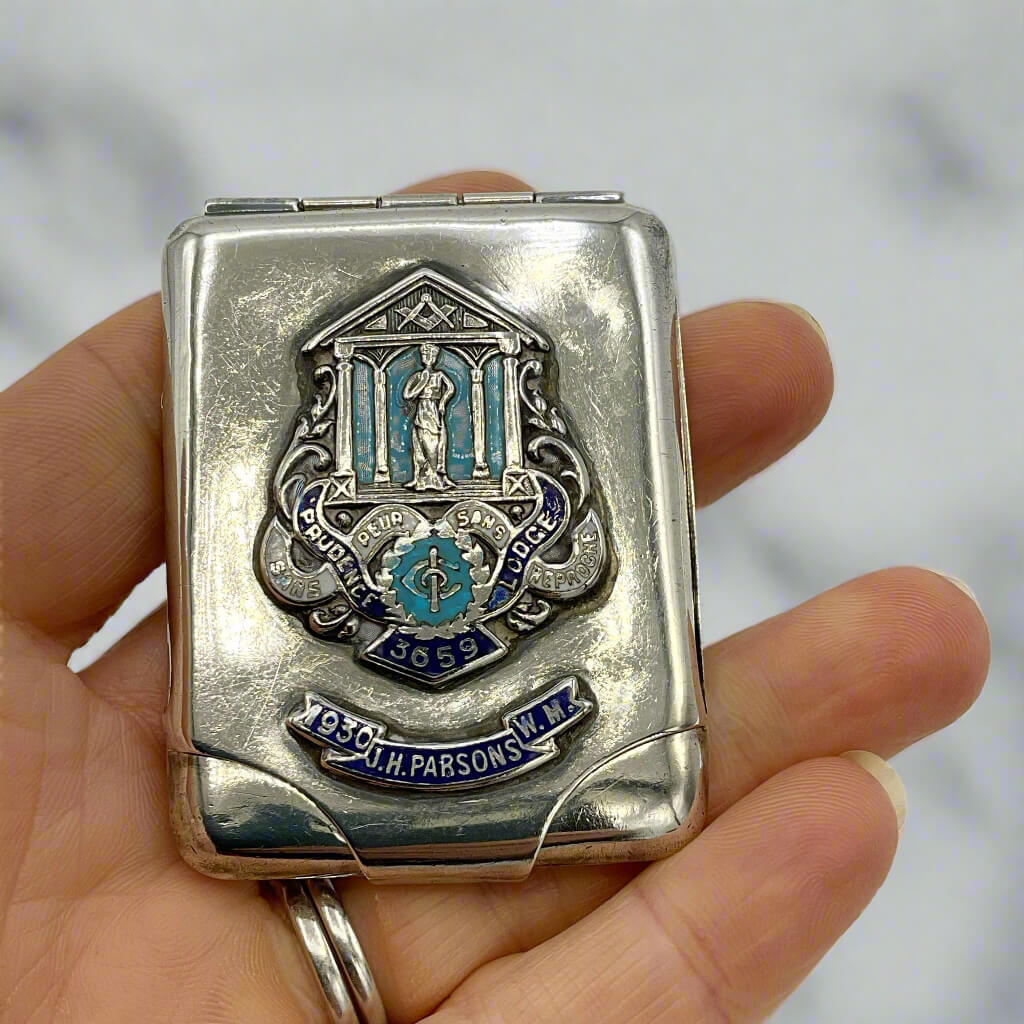 Silver matchbook case with decorative Masonic emblem held in a hand against a blurred background