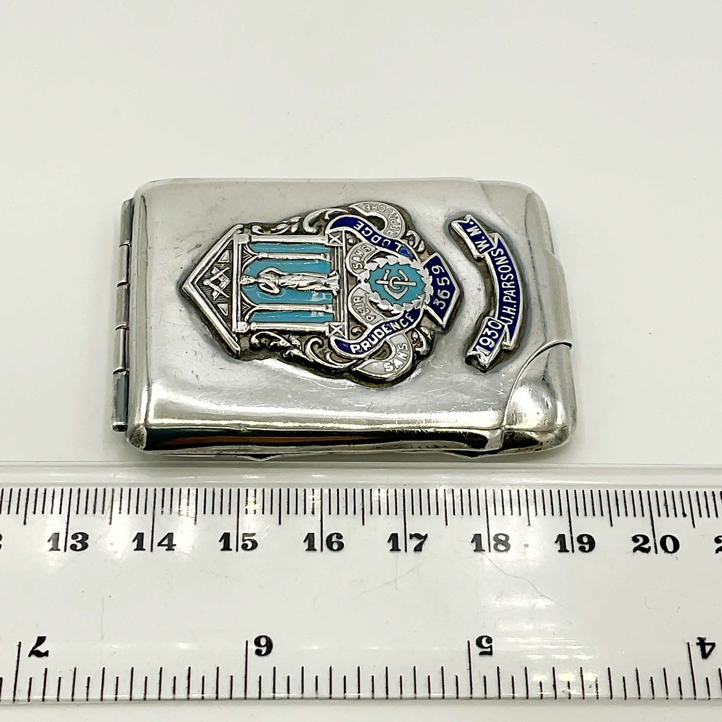 Silver matchbook with emblem and text on a white background, measured against a ruler.