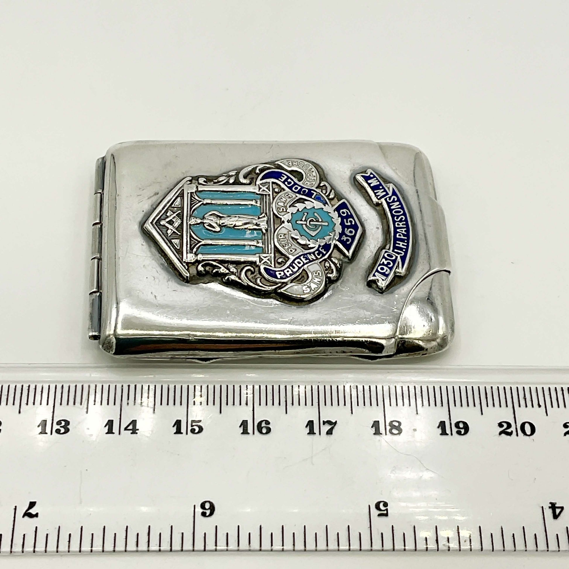 Silver matchbook with emblem and text on a white background, measured against a ruler.