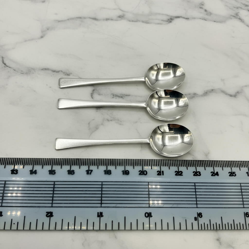 Three silver salt spoons on a marble surface with a ruler for scale.