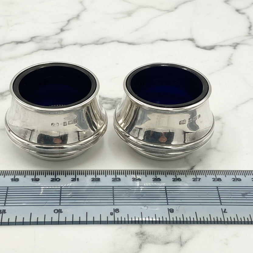 Two silver salts with blue glass liners next to a ruler for scale on a marble surface