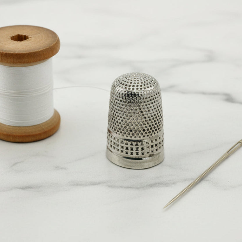 Silver thimble with wooden spool of thread and needle on a white marble background