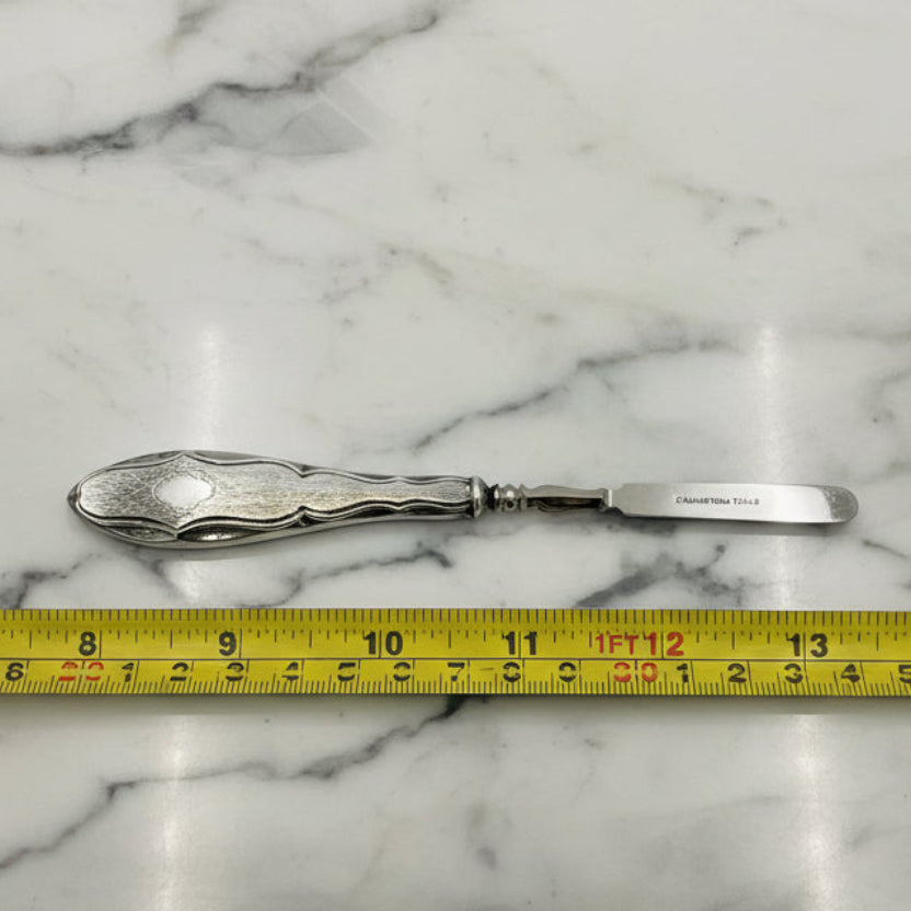 Silver cuticle pusher with decorative handle on a marble surface, measured by a yellow tape measure.