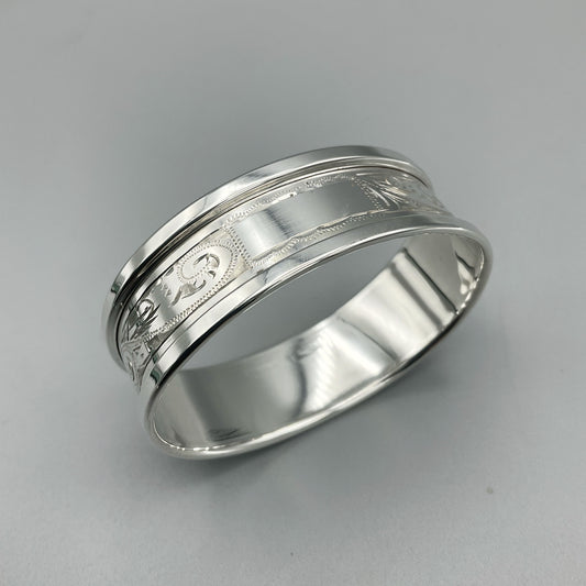 Silver napkin ring with blank rectangular cartouche and etched flower pattern sitting on a grey surface