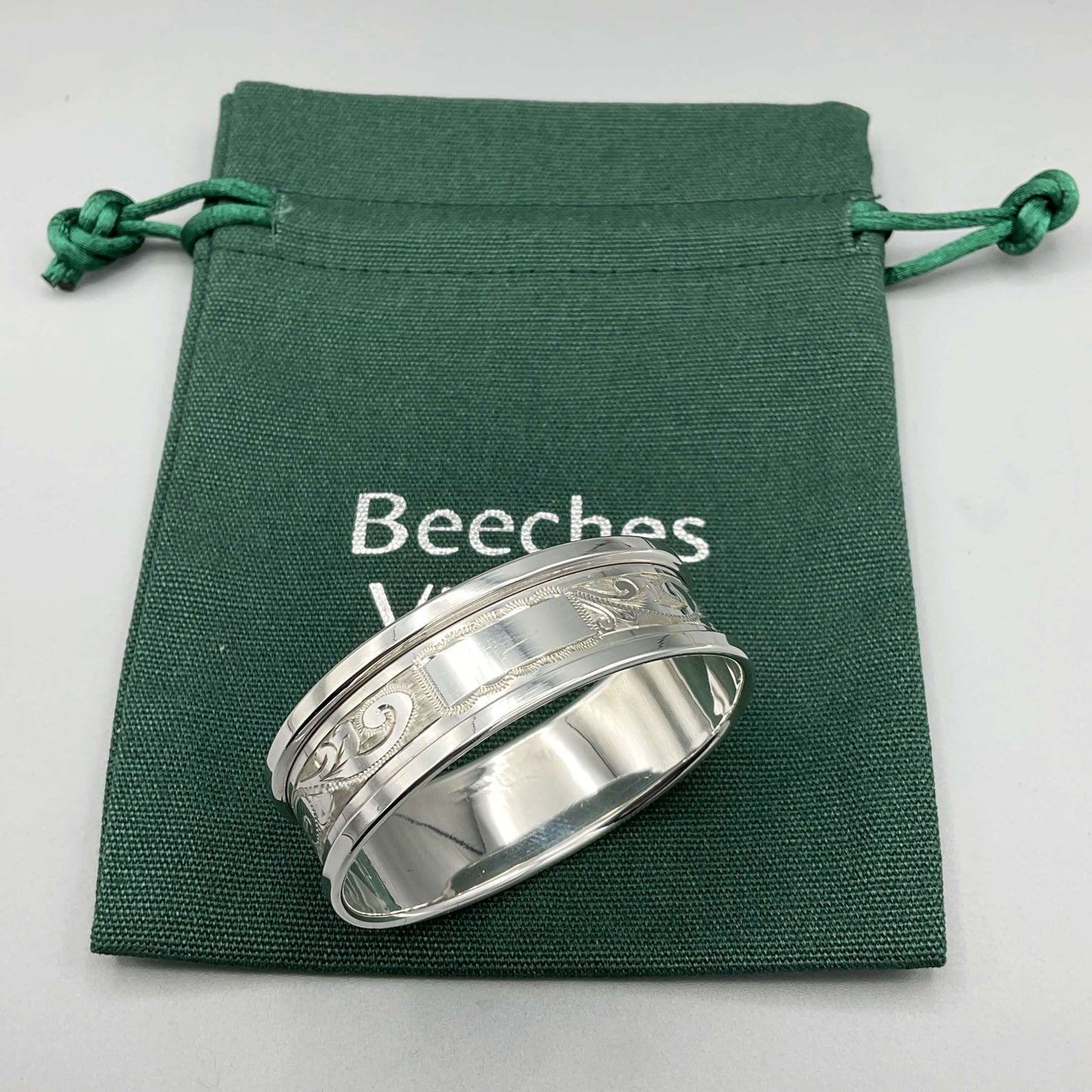 Silver napkin ring with blank rectangular cartouche and etched flower pattern sitting on a green bag