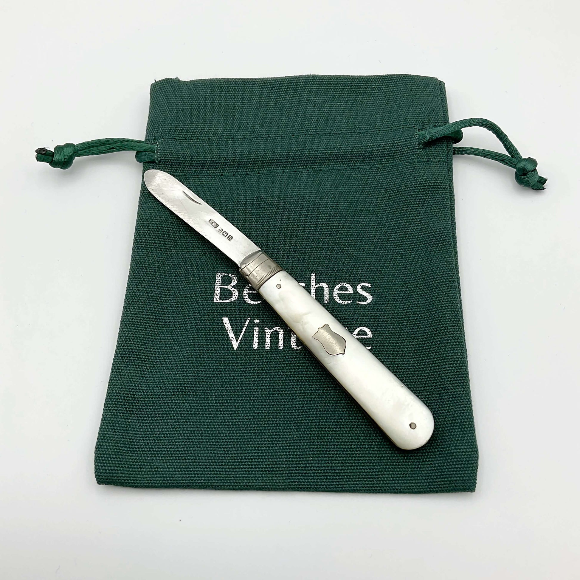 This is s folding fruit knife with a silver blade and a mother of pearl handle. It has a blank shield on one side of the handle. It's on a gift bag