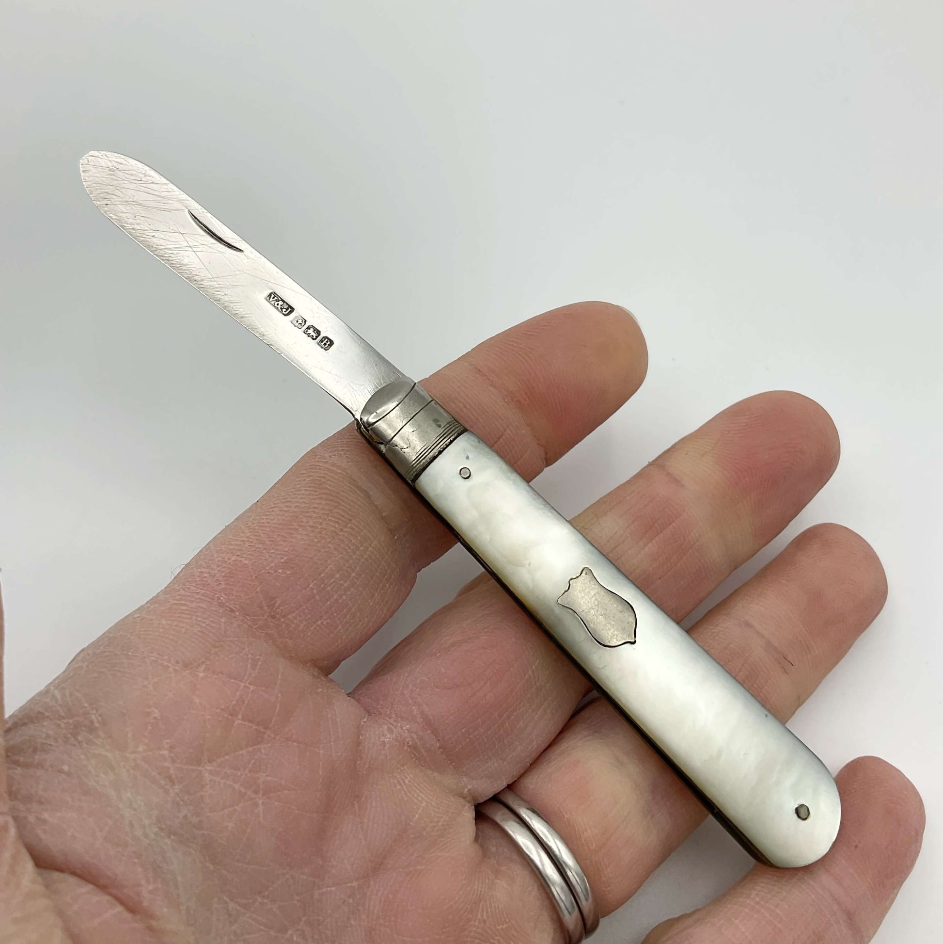 This is s folding fruit knife with a silver blade and a mother of pearl handle. It has a blank shield on one side of the handle held in a hand