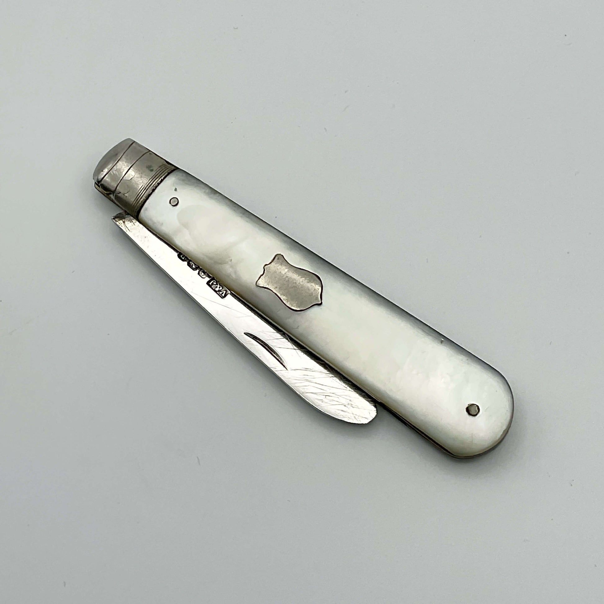 This is s folding fruit knife with a silver blade and a mother of pearl handle. It has a blank shield on one side of the handle with the blade folded in.