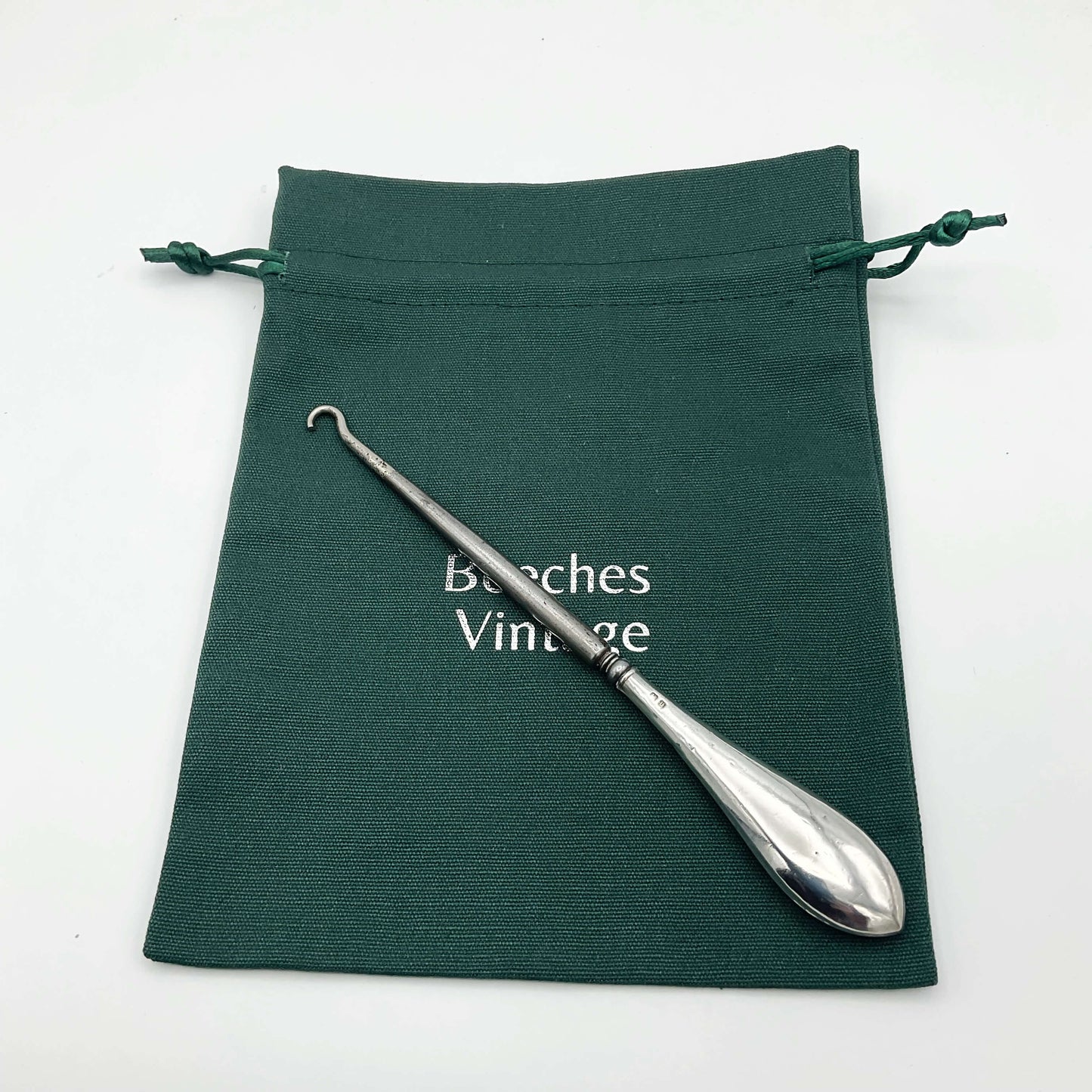 Silver handled button hook with a shiny handle and steel hook on a green cotton bag