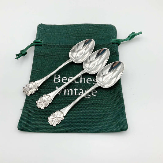 Three silver coffee spoons sitting on a green cotton bag with Beeches Vintage on it. Each spoon has a handle with a design featuring golf clubs and the initials BLGC. The bowls on the spoons have March 1932, April 1930 and June 1930 engraved on them.