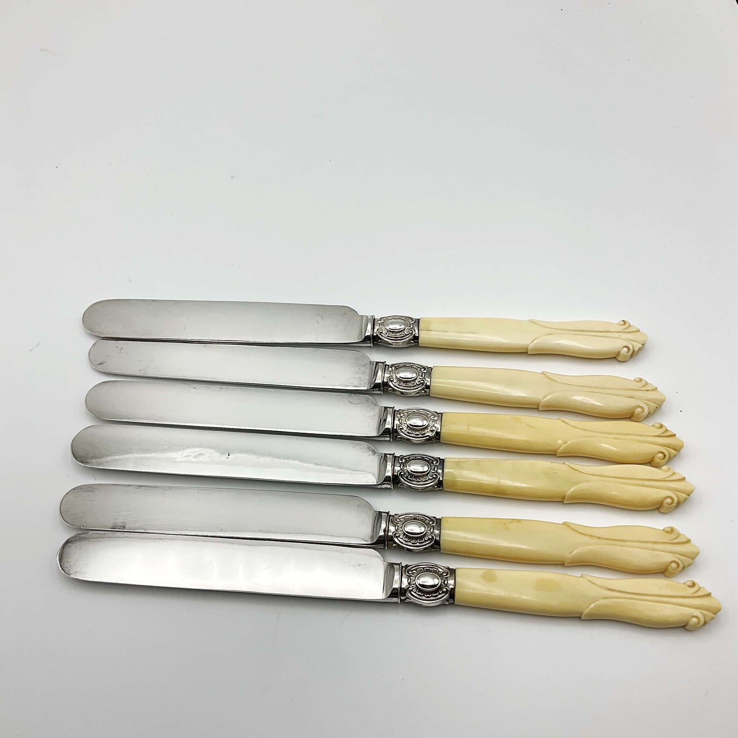 row of six silver plated bladed dessert knives with bone handles on a white background