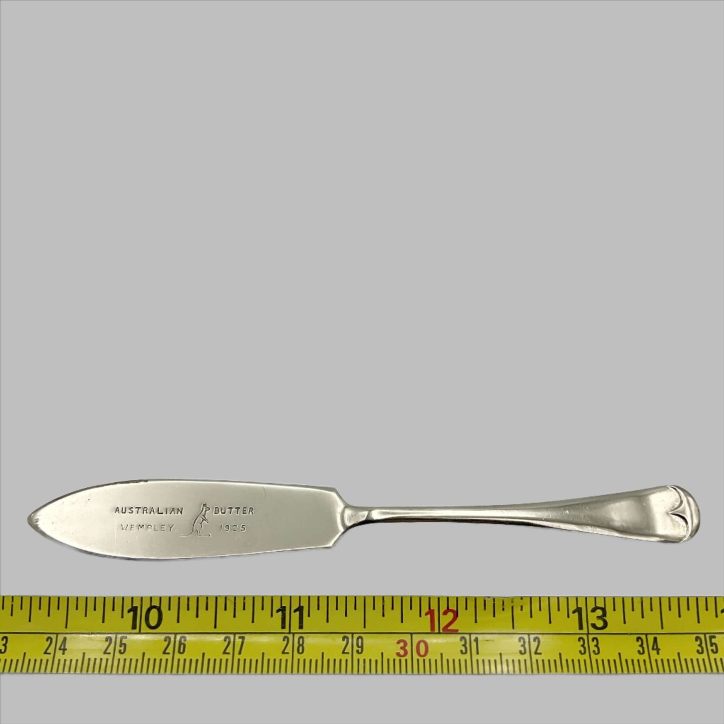Silver plated butter knife with Australian Butter Wembley 1925 on the blade with a Kangaroo emblem next to a tape measure