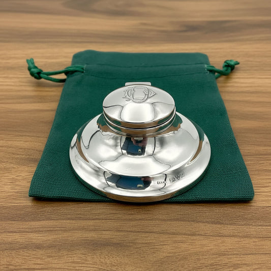 Antique sterling silver inkwell with a capstan shape, made in 1921, displayed on a green fabric pouch on a wood table.
