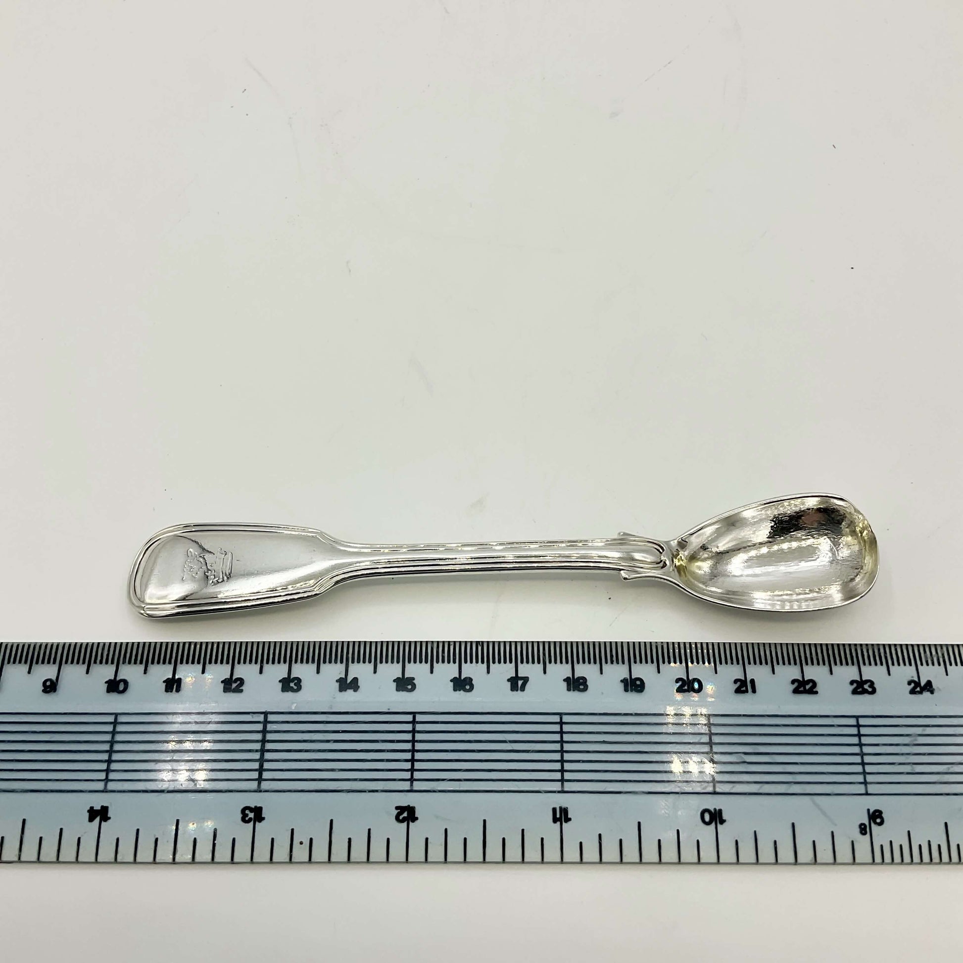 Antique silver mustard spoon held in a hand on a white background next to a ruler showing the length as 13.5cm