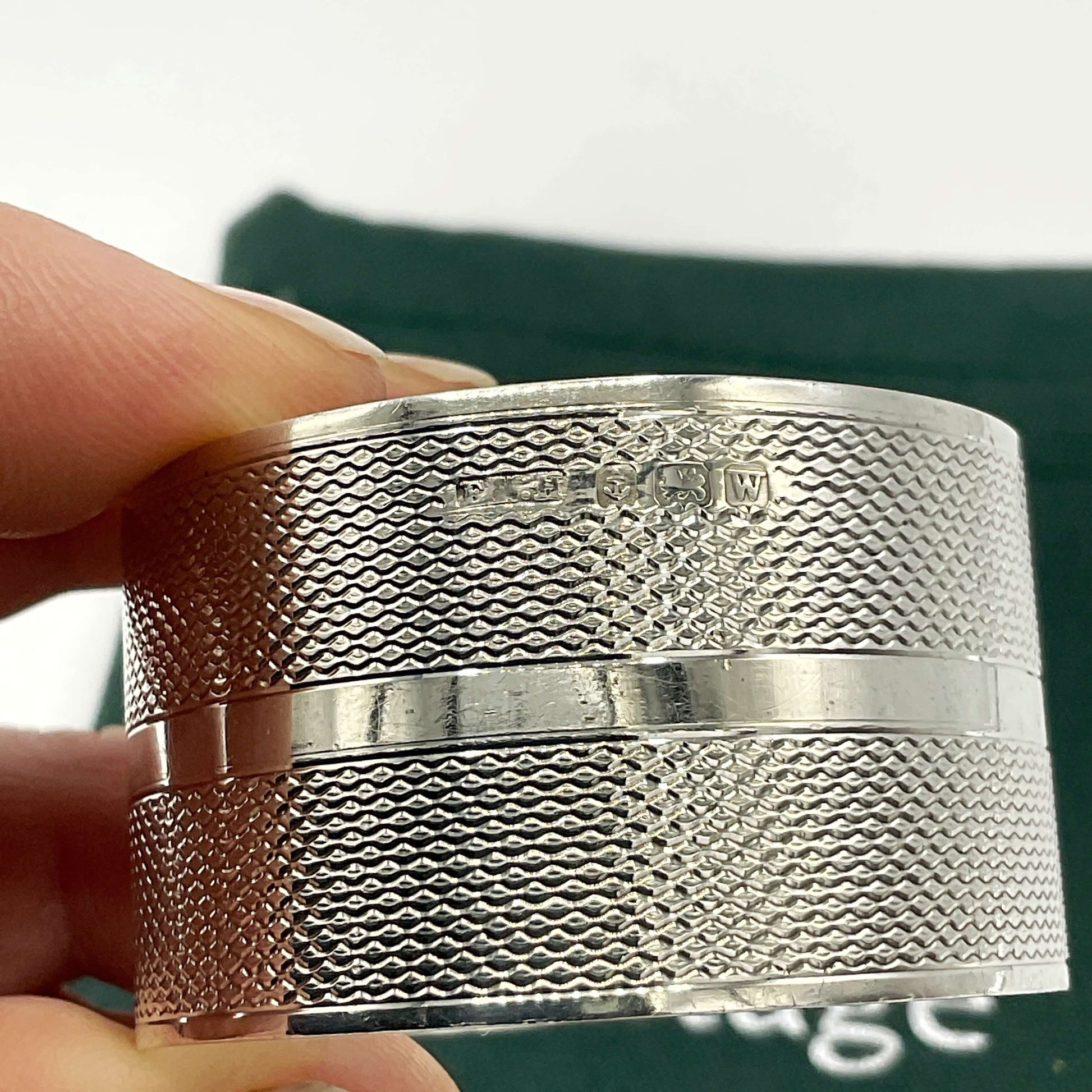 The maker’s mark and hallmarks on the side of a silver napkin ring held in fingers with a green gift bag in the background 