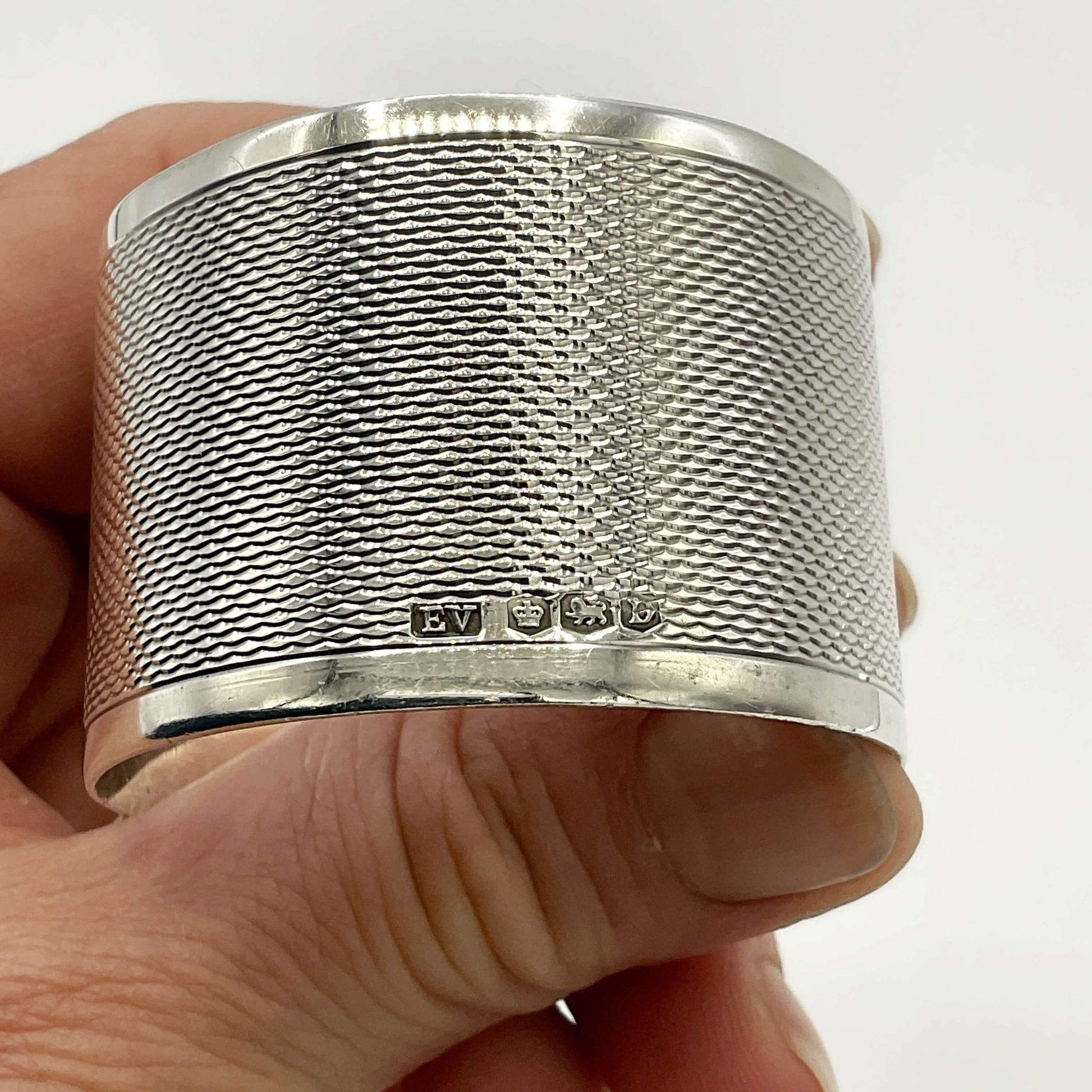 Silver napkin ring with the maker’s mark and hallmarks showing, held between fingers with a white background 