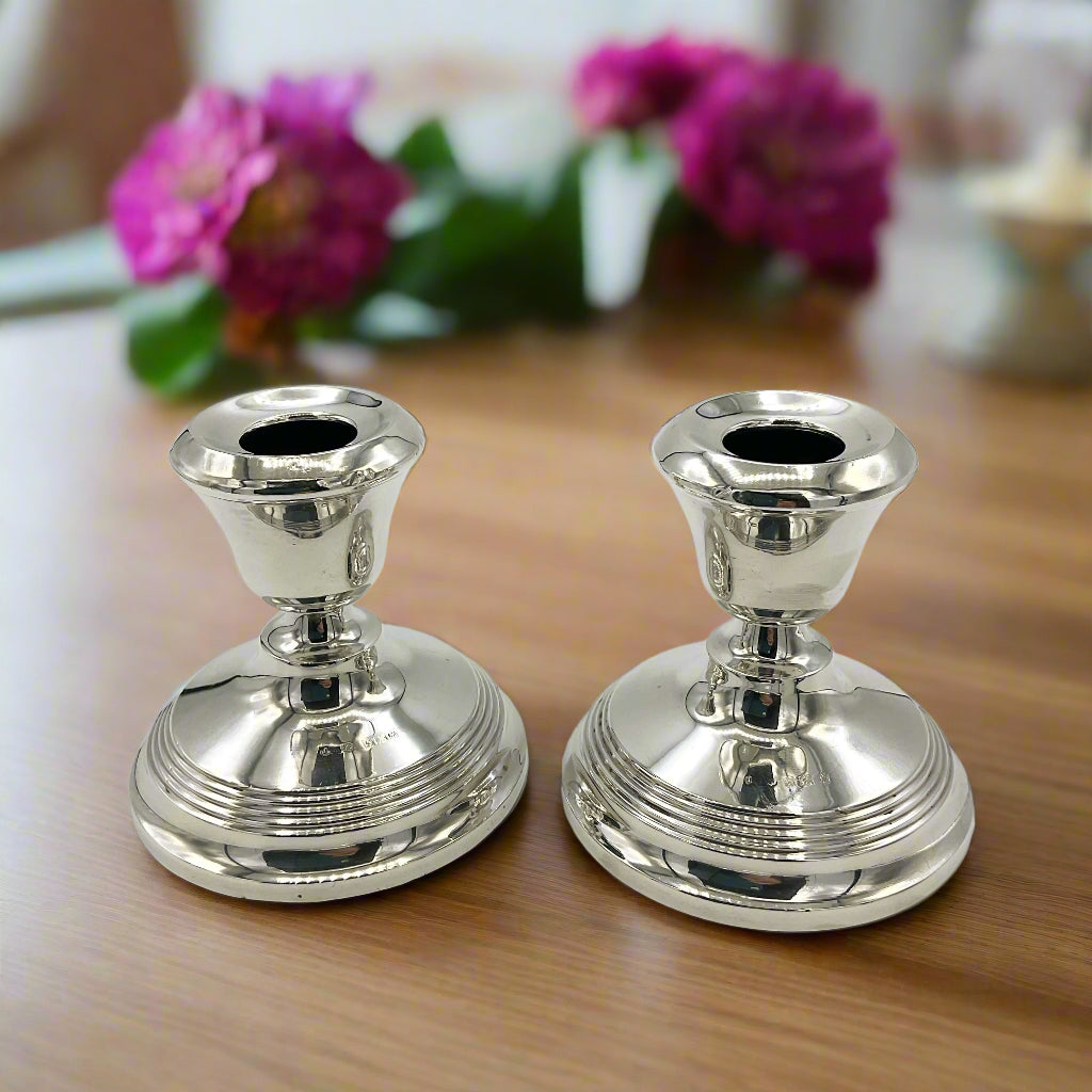 Two silver candle holders on a wooden table with flowers in the background 