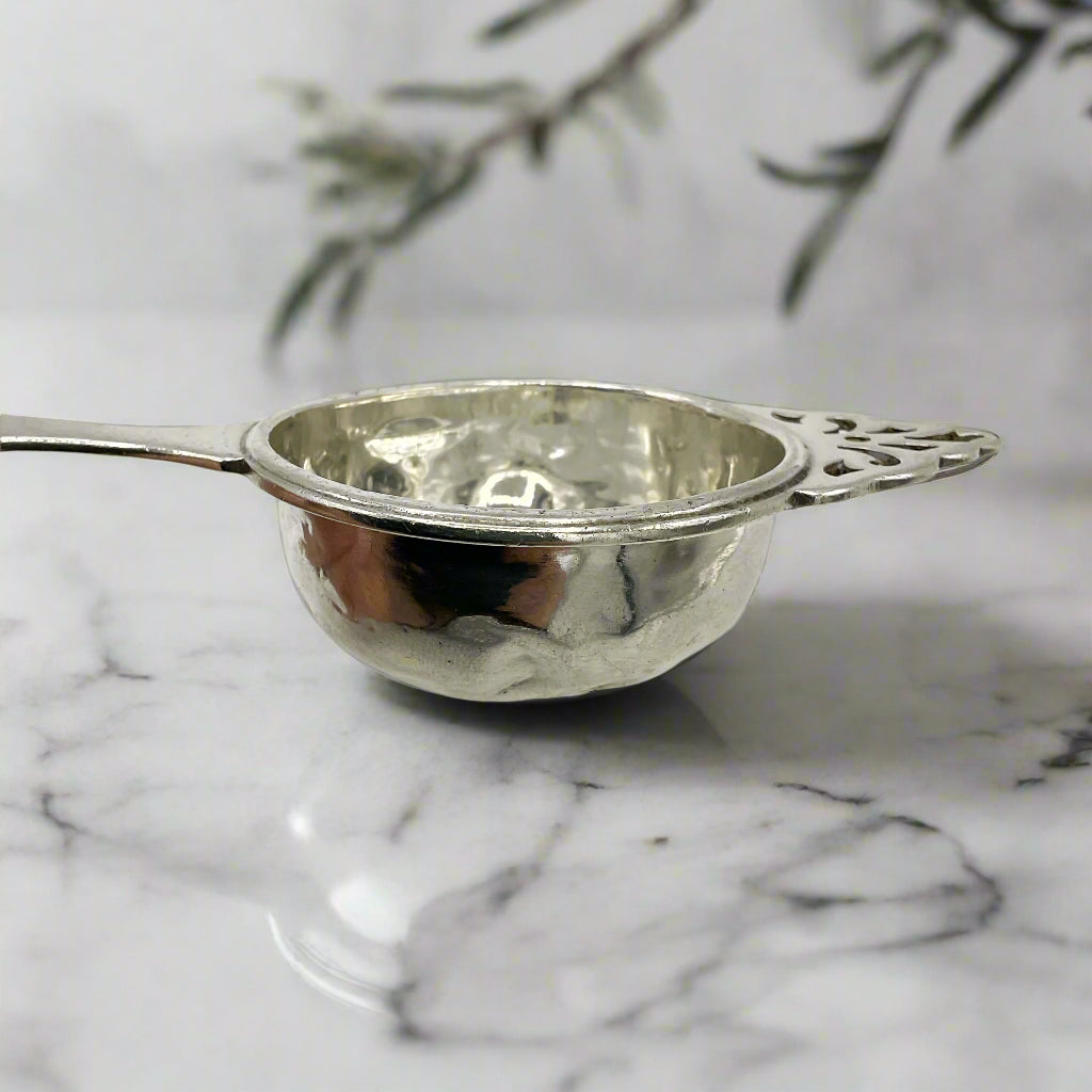 Side of silver tea strainer bowl with a marble worktop background and plant leaves
