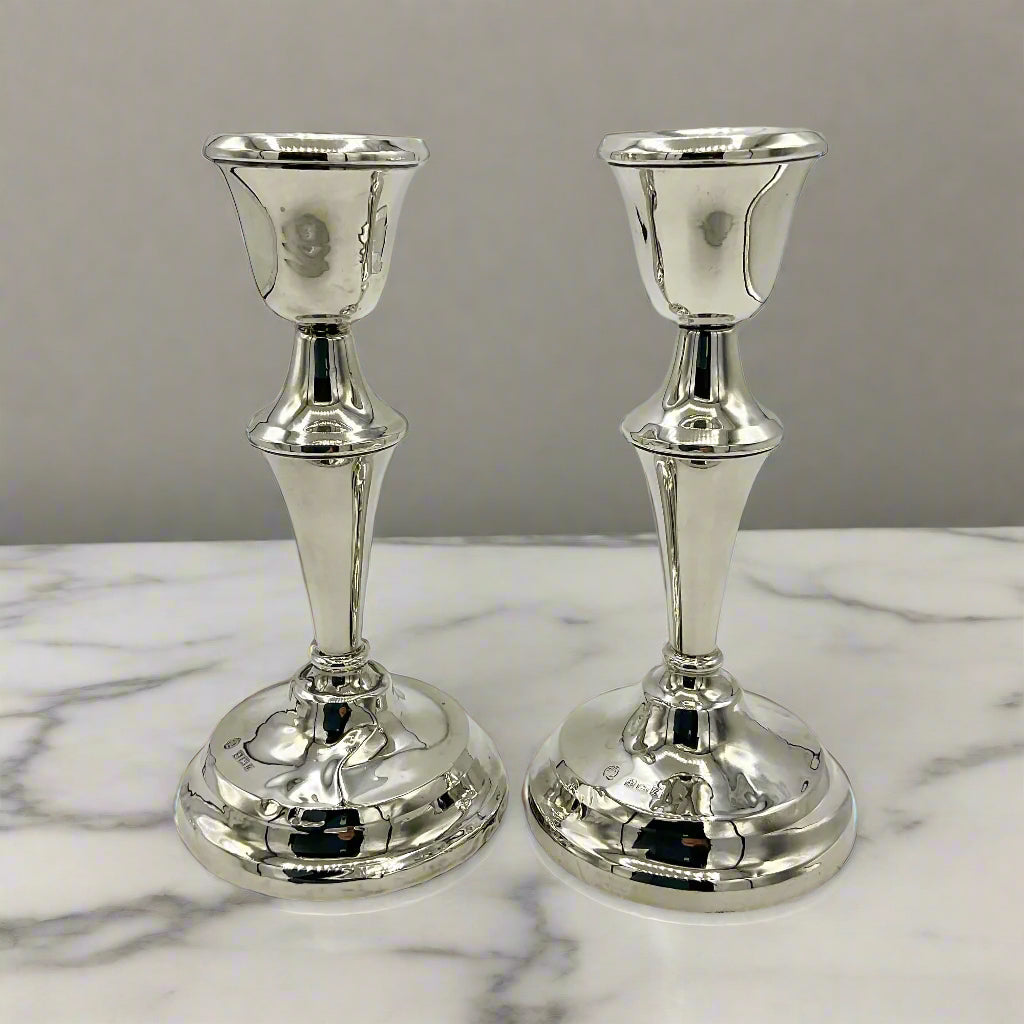 Two silver candle holders on a marble mantelpiece with a grey wall behind 
