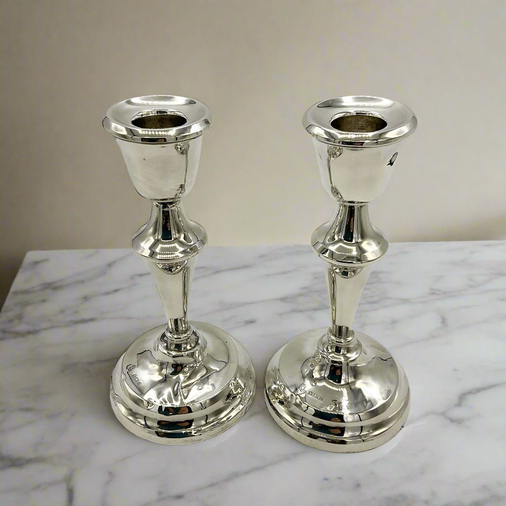 A matching pair of silver candlesticks on a marble mantelpiece with a cream wall in the background 