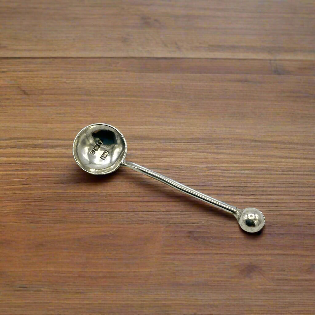 Antique 1902 small sterling silver salt spoon with ornate details, crafted by Beeches Vintage, resting on a reflective surface.