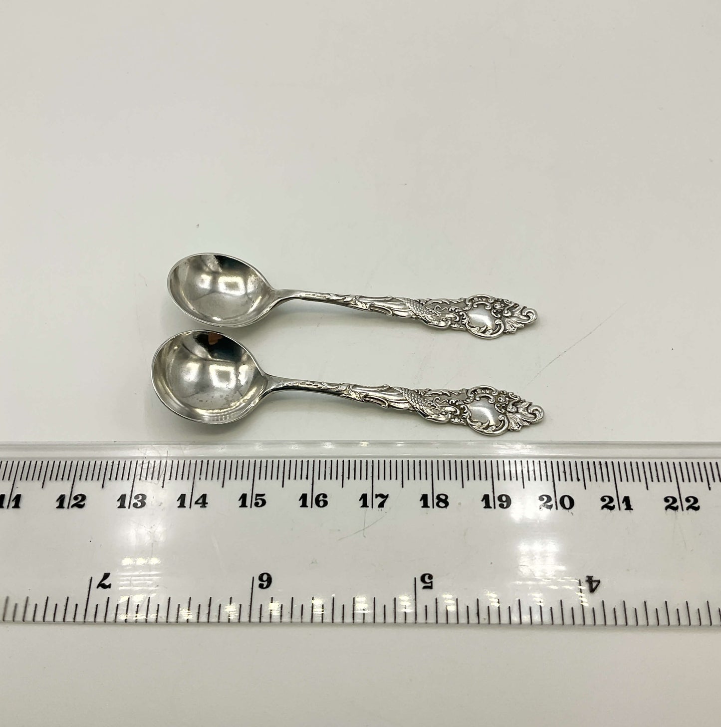 Two silver spoons on a white surface with a ruler for scale.