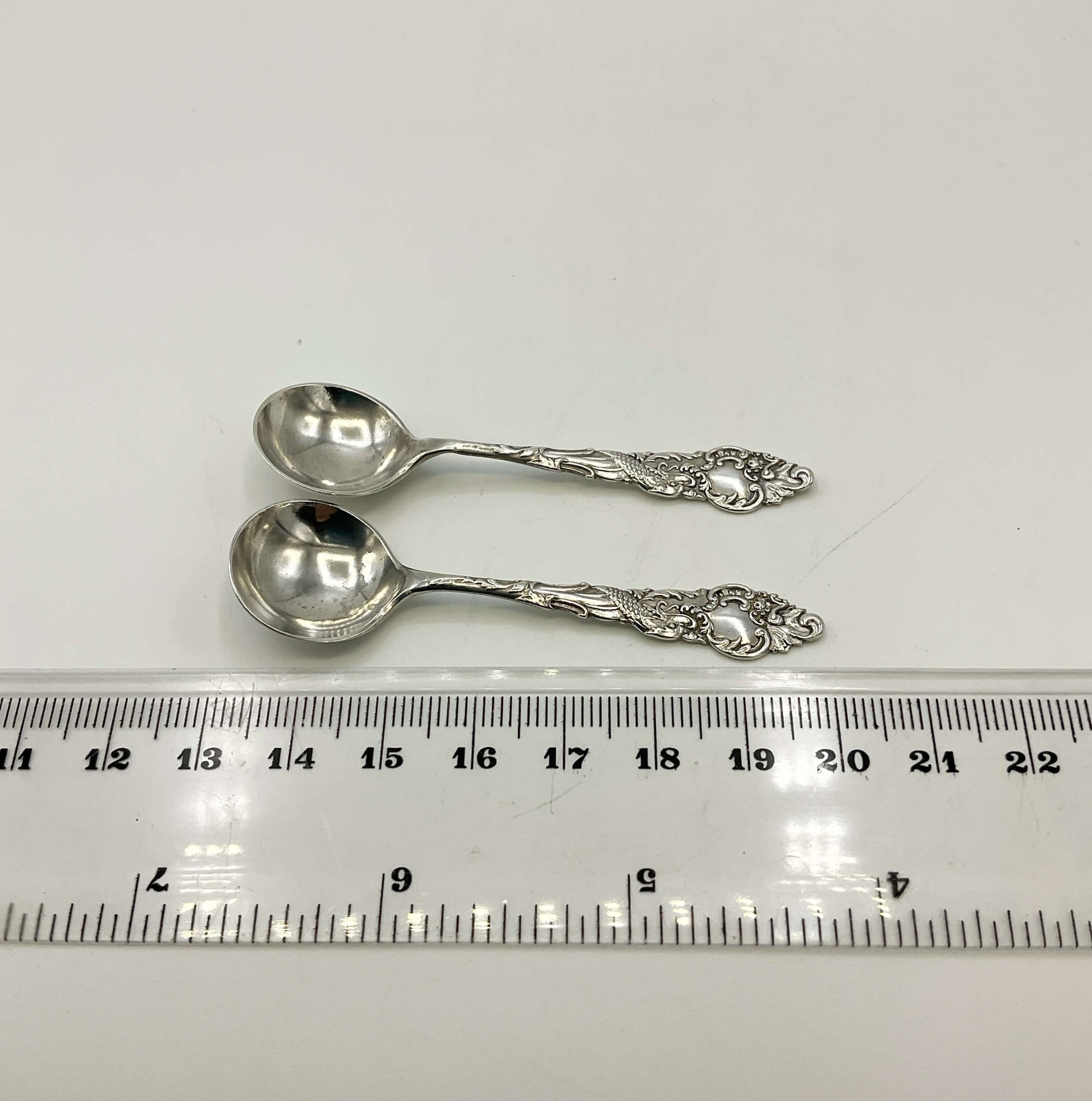 Two silver spoons on a white surface with a ruler for scale.