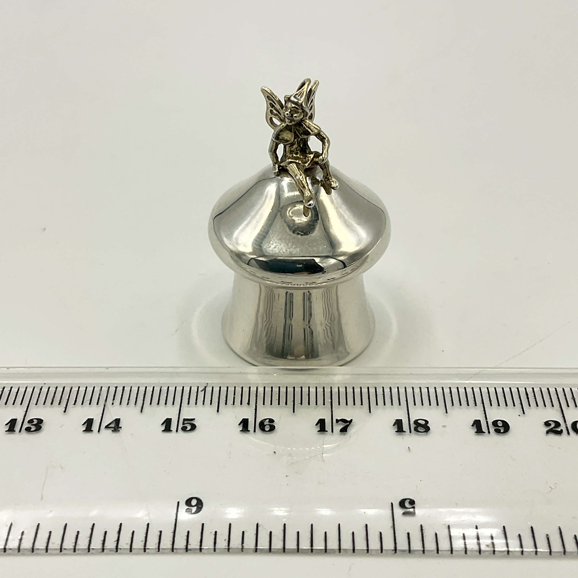 Silver pill box with a fairy on top on a white background with a ruler for scale.
