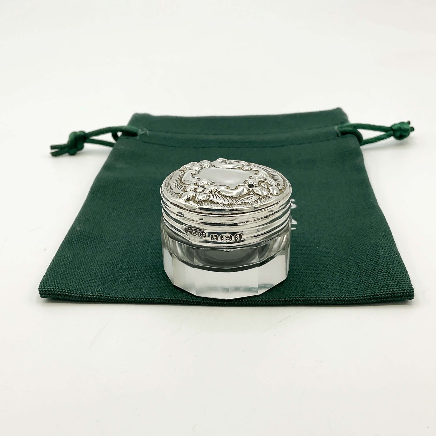 Decorative glass jar with silver lid on a green drawstring bag and a white background 