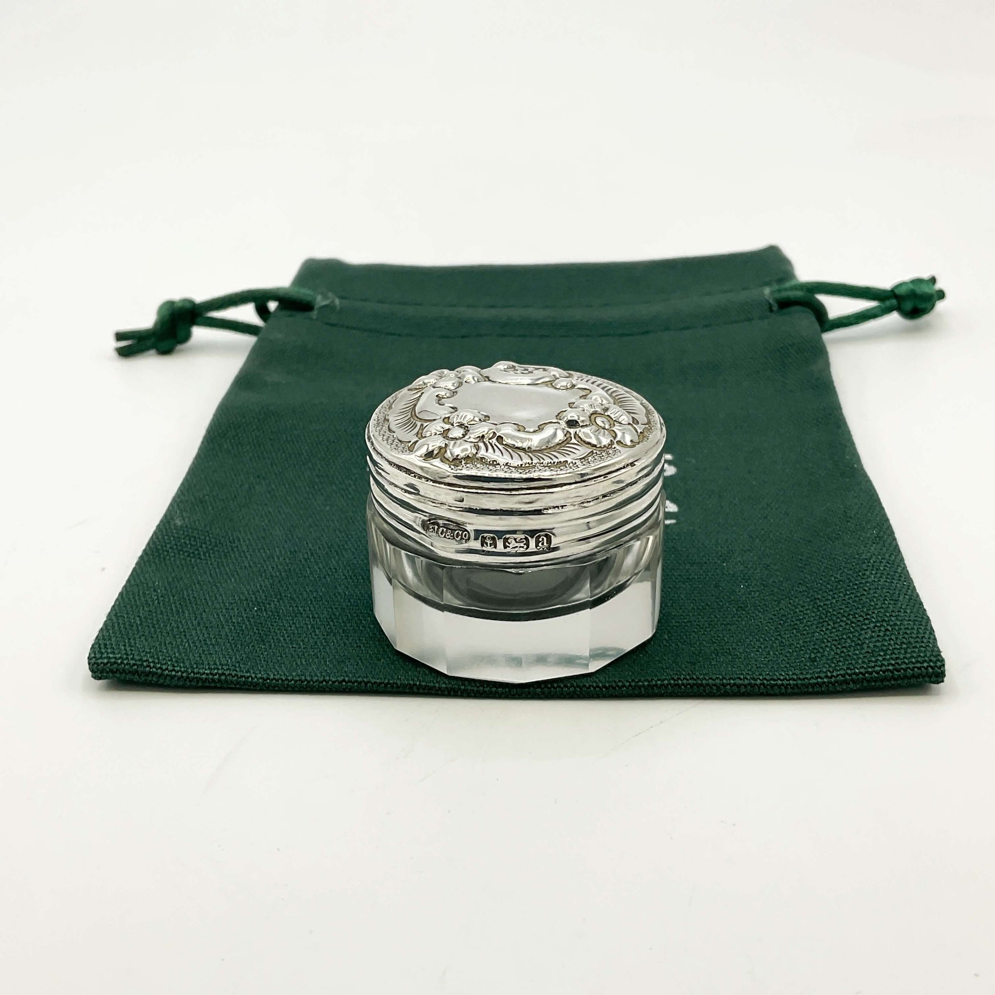 Decorative glass jar with silver lid on a green drawstring bag and a white background 