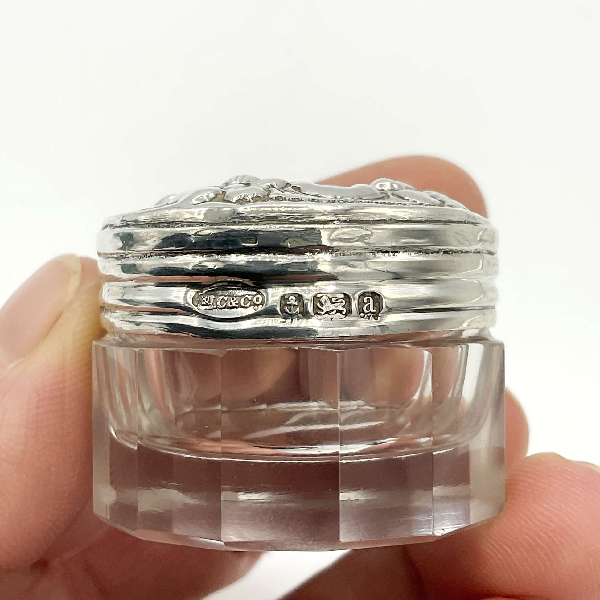 Small glass jar with silver lid showing hallmarks held between fingers against a white background