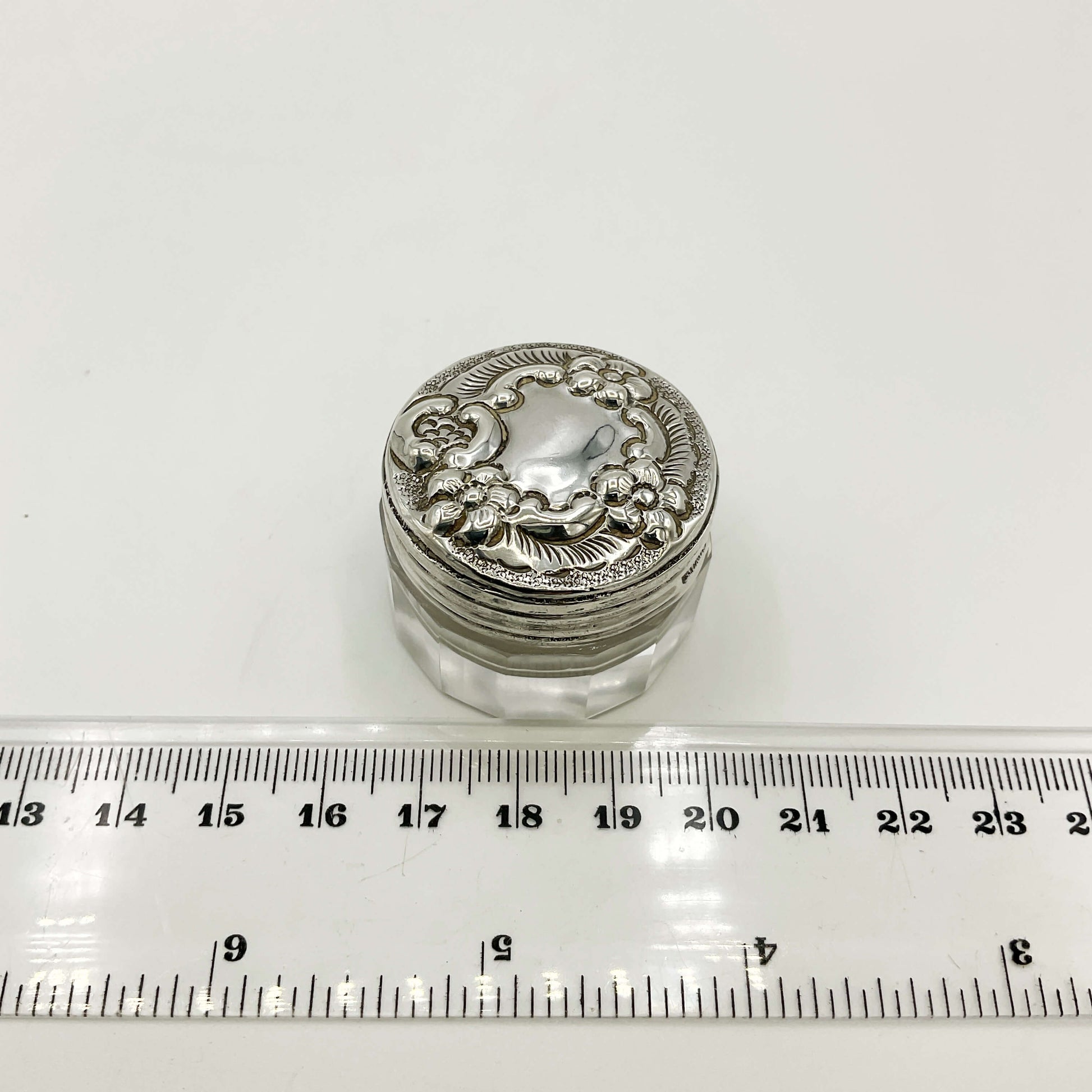 Decorative silver lid on a glass container with a ruler for scale on a white background