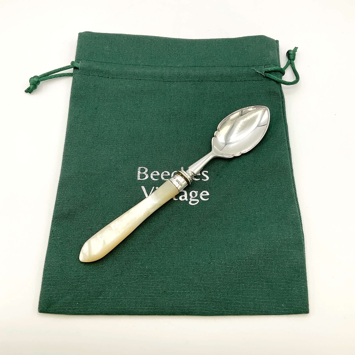 Silver spoon with a white handle on a green drawstring bag with 'Beeches Vintage' branding.